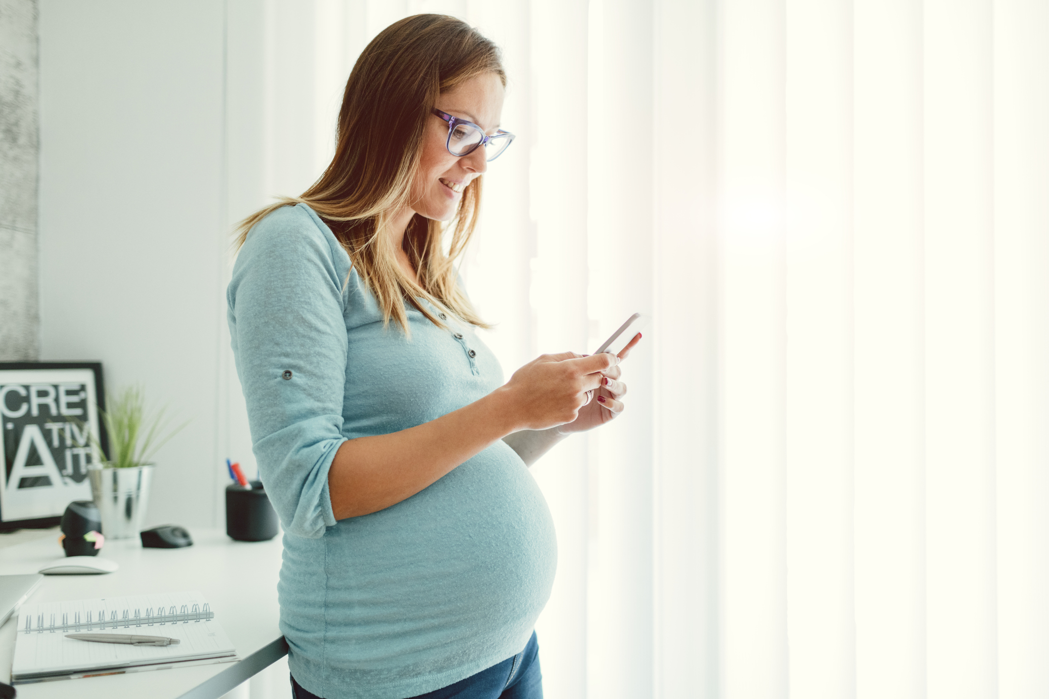 Pregnant Businesswoman Texting On Smart Phone In Her Home Office.